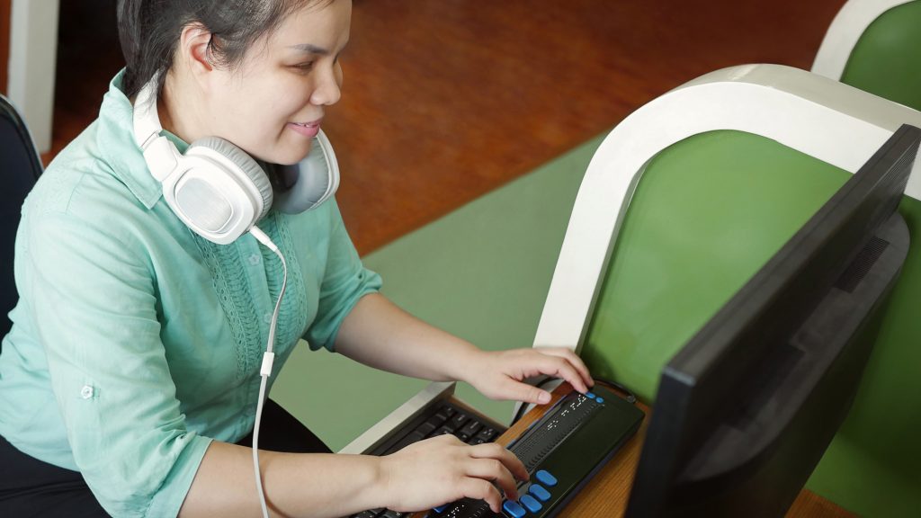 Asian young blind woman with headphone using computer with refreshable braille display or braille terminal a technology device for persons with visual disabilities.